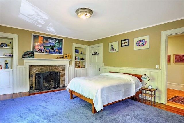 bedroom with crown molding, a wainscoted wall, a brick fireplace, and wood finished floors