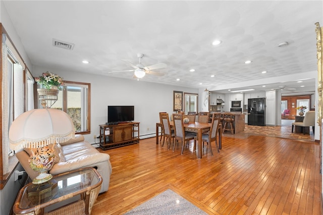 living area with visible vents, ceiling fan, a baseboard heating unit, light wood finished floors, and a wealth of natural light