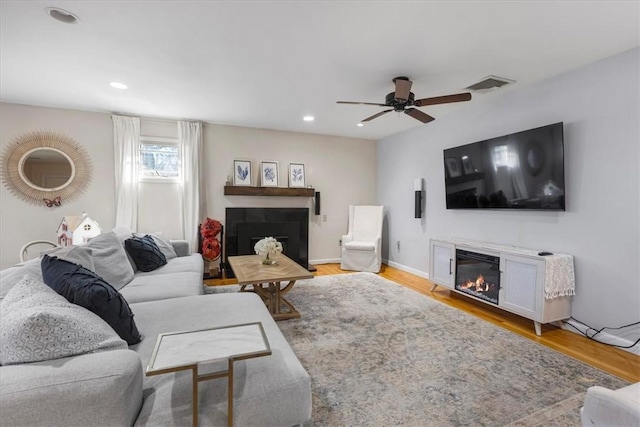 living area featuring a ceiling fan, a glass covered fireplace, wood finished floors, and visible vents