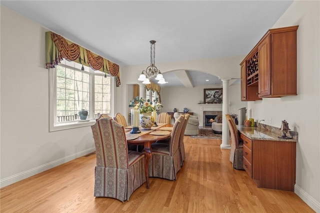 dining space featuring baseboards, a lit fireplace, ornate columns, arched walkways, and light wood finished floors