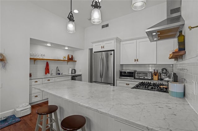 kitchen featuring stainless steel appliances, wall chimney range hood, white cabinetry, backsplash, and a breakfast bar