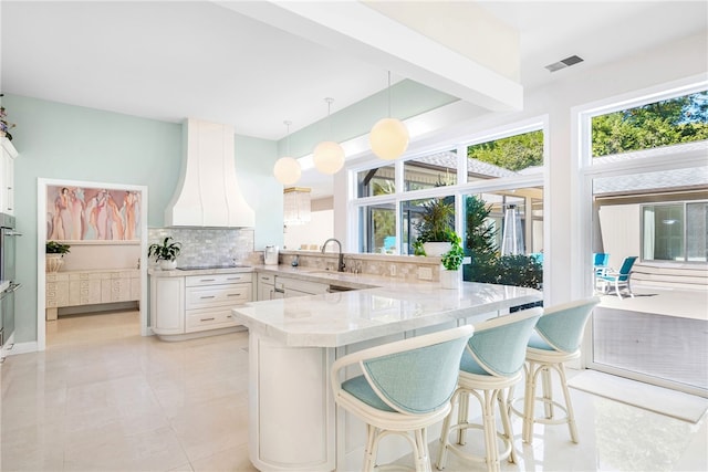 kitchen featuring a breakfast bar area, a sink, premium range hood, a peninsula, and decorative backsplash