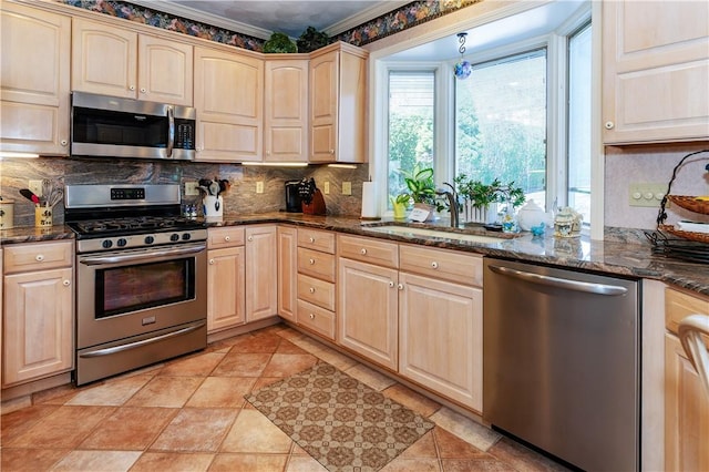 kitchen featuring appliances with stainless steel finishes, light brown cabinetry, a sink, decorative backsplash, and dark stone counters
