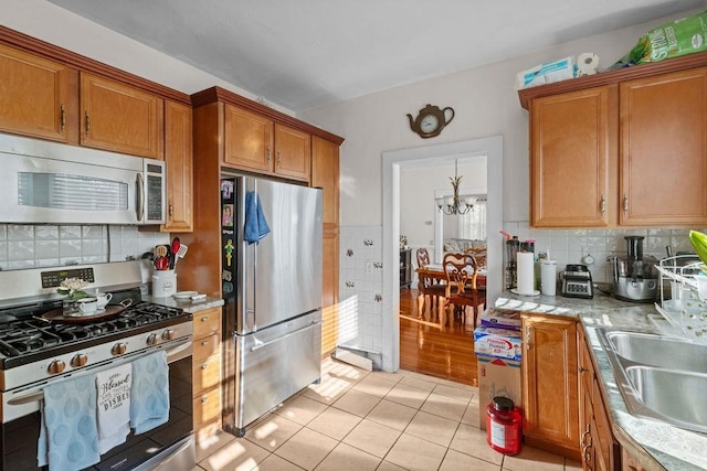 kitchen with brown cabinetry, stainless steel appliances, tasteful backsplash, light tile patterned floors, and a notable chandelier