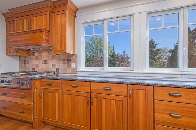 kitchen with brown cabinetry, tasteful backsplash, dark stone countertops, stainless steel gas cooktop, and light wood finished floors