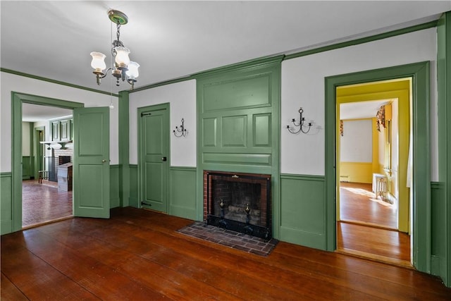 unfurnished living room featuring wainscoting, a brick fireplace, a notable chandelier, and dark wood-type flooring