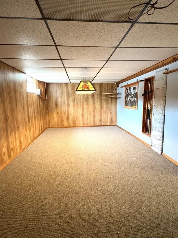 below grade area featuring a paneled ceiling, wooden walls, carpet, and baseboards