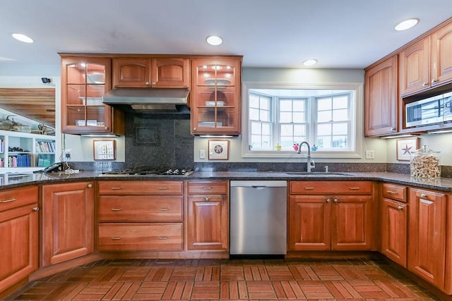 kitchen with stainless steel appliances, under cabinet range hood, a sink, dark stone countertops, and glass insert cabinets