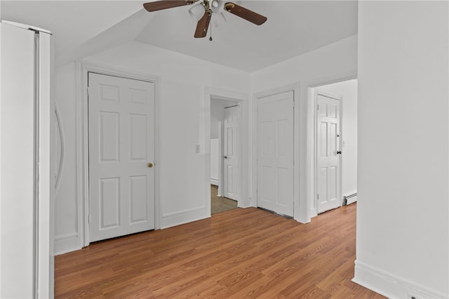 unfurnished bedroom featuring a baseboard radiator, freestanding refrigerator, light wood-type flooring, ceiling fan, and baseboards
