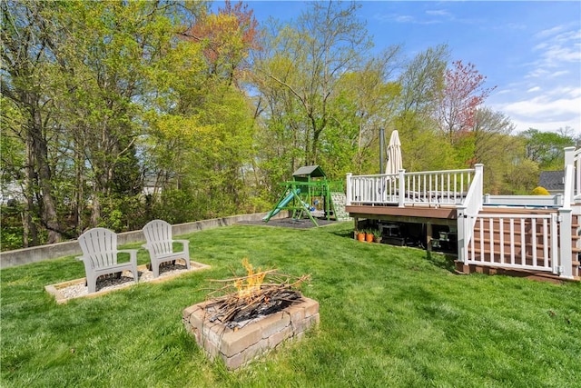 view of grassy yard with an outdoor fire pit, a playground, a deck, and view of scattered trees
