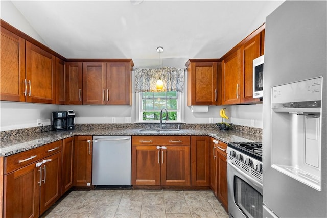 kitchen with a sink, stainless steel appliances, dark stone countertops, brown cabinetry, and hanging light fixtures