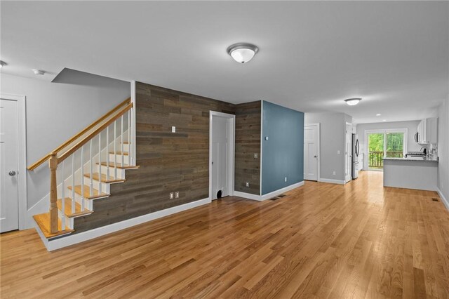unfurnished living room with stairs, an accent wall, light wood finished floors, baseboards, and wood walls