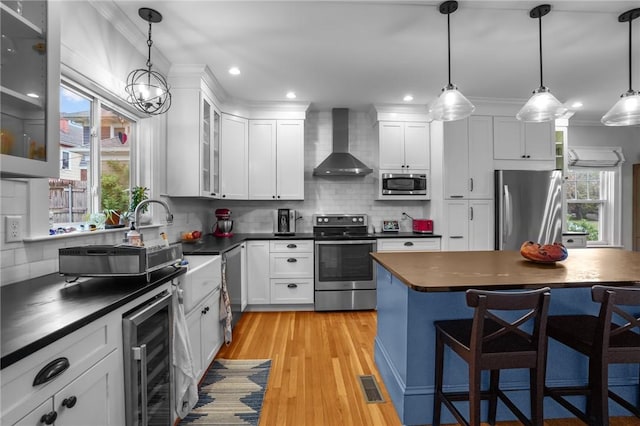kitchen with appliances with stainless steel finishes, wall chimney exhaust hood, beverage cooler, dark countertops, and a kitchen bar
