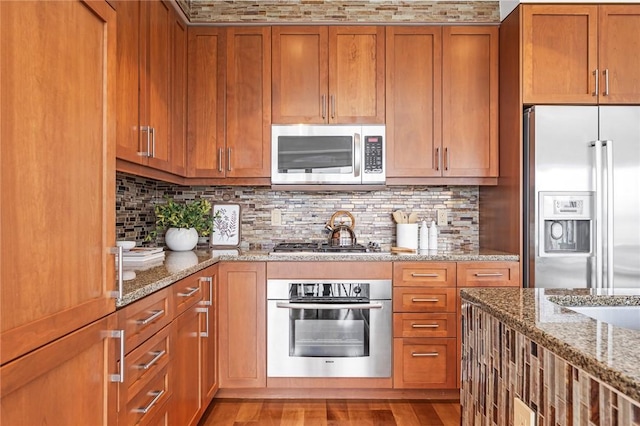 kitchen with stainless steel appliances, brown cabinets, backsplash, light stone countertops, and light wood-style floors