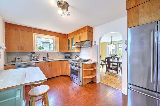 kitchen with stainless steel appliances, under cabinet range hood, a sink, arched walkways, and dark wood-type flooring