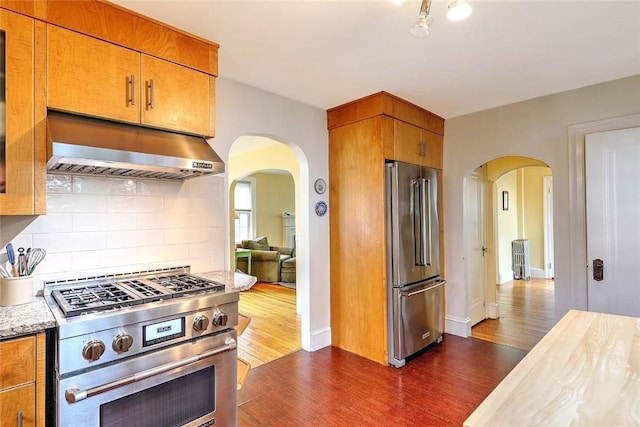 kitchen featuring high quality appliances, arched walkways, dark wood-style flooring, under cabinet range hood, and backsplash
