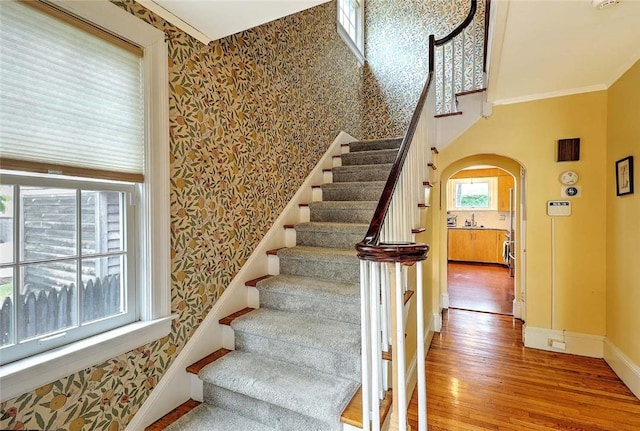 stairs with arched walkways, wood finished floors, wallpapered walls, baseboards, and ornamental molding