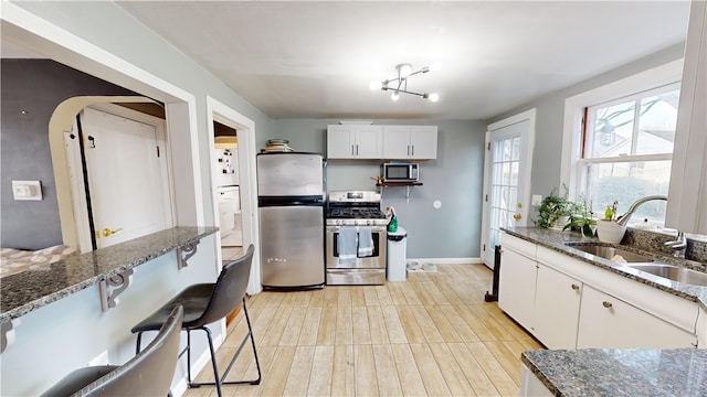 kitchen featuring stainless steel appliances, a sink, dark stone counters, white cabinetry, and baseboards