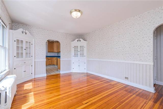 43 Standish Rd, Jamestown, RI 02835 - unfurnished living room featuring arched walkways, light wood-style flooring, wainscoting, wallpapered walls, and radiator heating unit