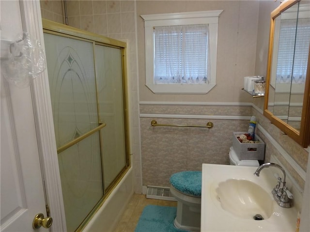 full bath featuring toilet, tile walls, tile patterned flooring, combined bath / shower with glass door, and a wainscoted wall