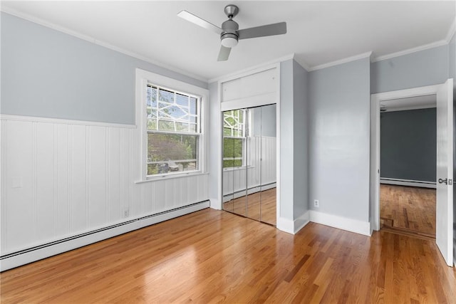 unfurnished bedroom with a baseboard heating unit, wood finished floors, crown molding, wainscoting, and ceiling fan