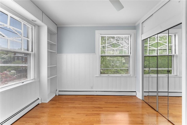 spare room featuring baseboard heating, light wood-type flooring, healthy amount of natural light, a wainscoted wall, and crown molding