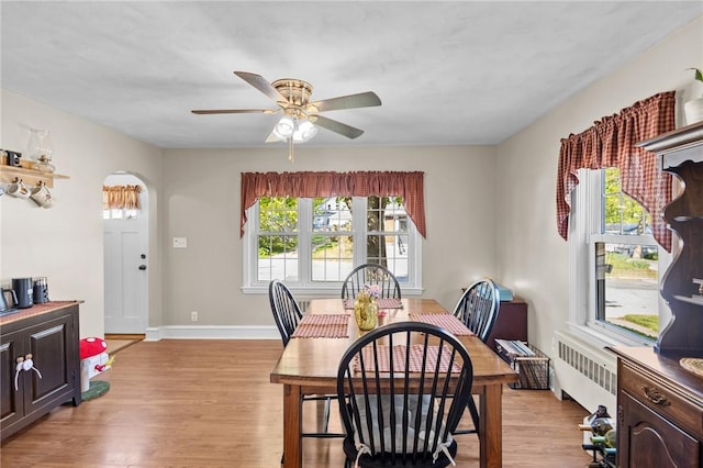 dining area with radiator, light wood-style floors, ceiling fan, arched walkways, and baseboards