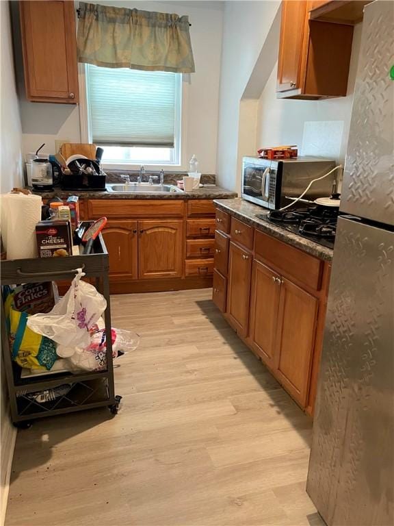kitchen featuring a sink, appliances with stainless steel finishes, light wood-style flooring, dark countertops, and brown cabinets