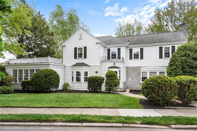 view of front of property featuring a chimney, a front yard, stone siding, a shingled roof, and a balcony