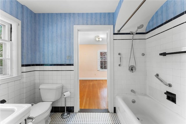 bathroom with wallpapered walls, toilet, shower / bathtub combination, wainscoting, and tile walls