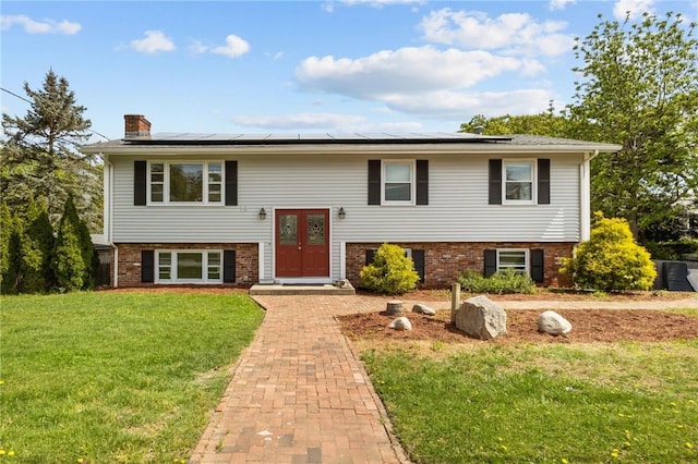 raised ranch featuring a chimney, a front yard, brick siding, and solar panels