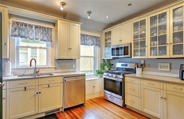 kitchen featuring stainless steel appliances, a sink, light wood-type flooring, glass insert cabinets, and light stone countertops