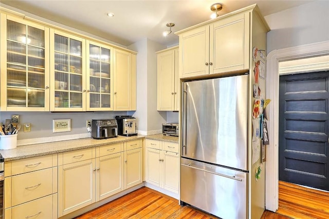 kitchen with freestanding refrigerator, light wood-type flooring, glass insert cabinets, light stone countertops, and cream cabinetry