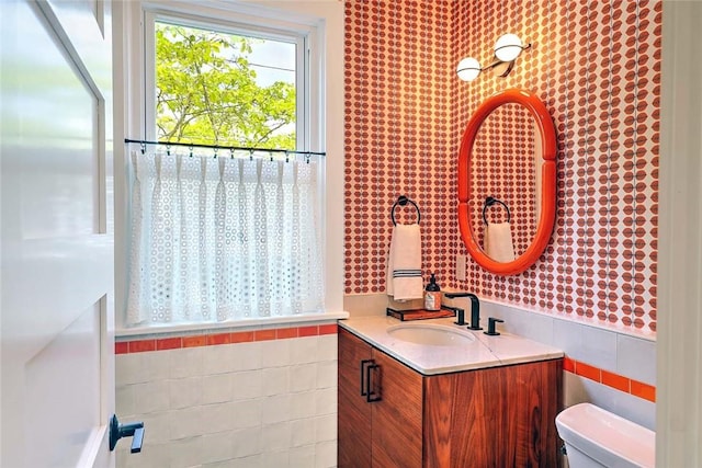 bathroom featuring tile walls, toilet, vanity, wallpapered walls, and a wainscoted wall