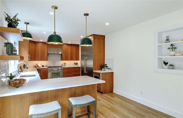 kitchen with a sink, high quality appliances, wall chimney range hood, brown cabinetry, and baseboards