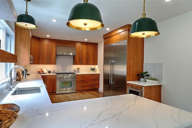 kitchen featuring premium appliances, a sink, brown cabinetry, exhaust hood, and recessed lighting