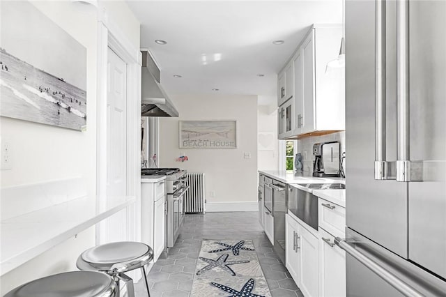 286 Kingstown Rd, Narragansett, RI 02882 - kitchen featuring premium appliances, wall chimney range hood, radiator, light countertops, and white cabinetry