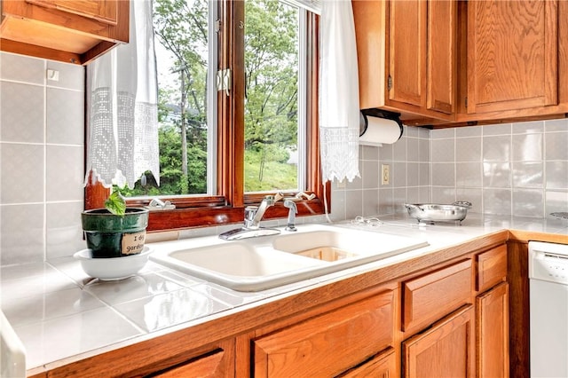 kitchen featuring white dishwasher, a sink, light countertops, decorative backsplash, and brown cabinetry