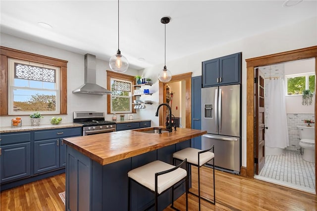 kitchen featuring appliances with stainless steel finishes, wall chimney exhaust hood, a sink, butcher block counters, and blue cabinetry