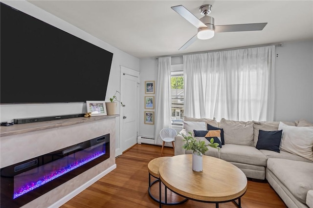 living area with a glass covered fireplace, wood finished floors, ceiling fan, and baseboard heating