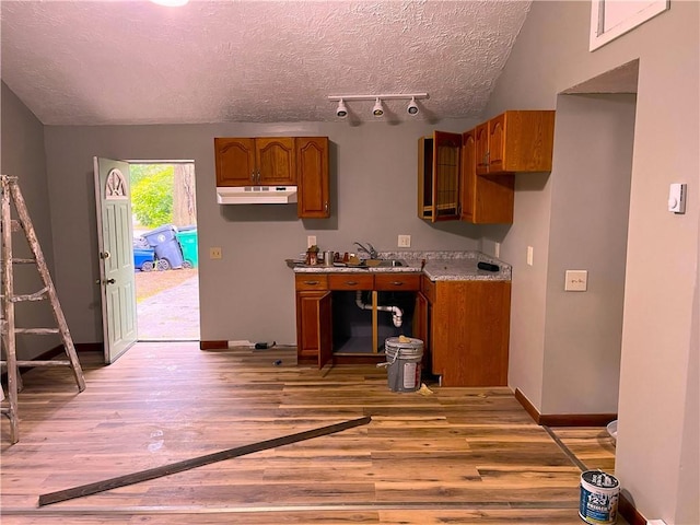 kitchen with brown cabinetry, light wood finished floors, a textured ceiling, baseboards, and under cabinet range hood