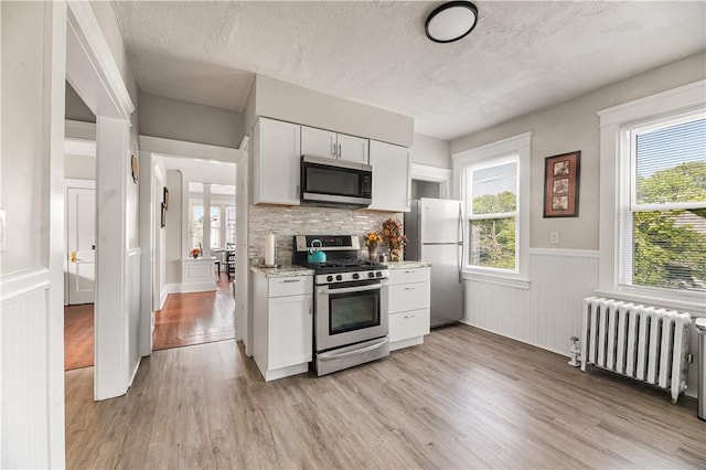 61 Wealth Ave, Providence, RI 02908 - kitchen featuring appliances with stainless steel finishes, radiator heating unit, a wainscoted wall, light wood finished floors, and white cabinetry