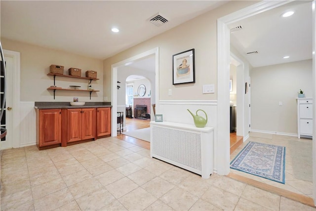 72 Audubon Rd, Warwick, RI 02888 - kitchen featuring arched walkways, dark countertops, a brick fireplace, brown cabinetry, and a wainscoted wall