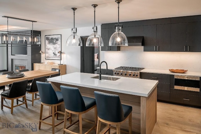 kitchen featuring pendant lighting, light countertops, light wood-style flooring, a kitchen island with sink, and a sink