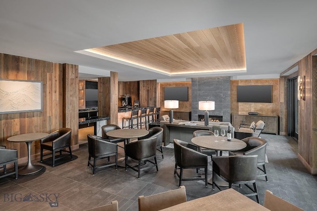 dining room with a tray ceiling, a tiled fireplace, wood ceiling, and wooden walls