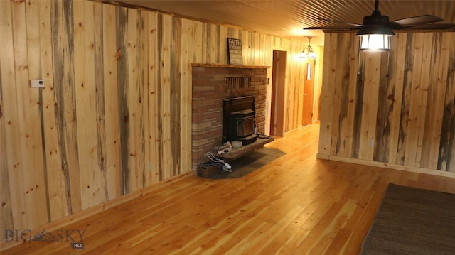 unfurnished living room with ceiling fan, a wood stove, wood-type flooring, wooden walls, and a fireplace