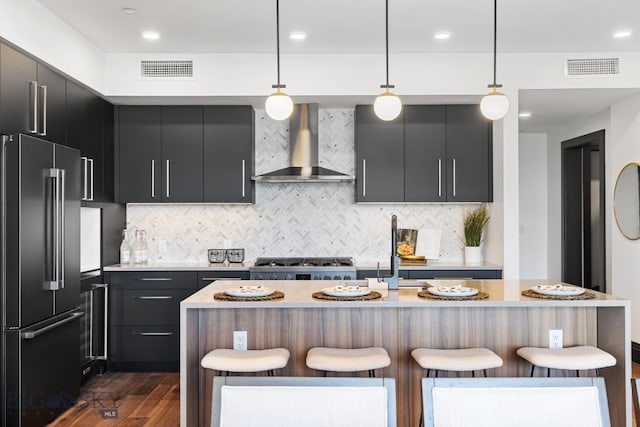 kitchen with appliances with stainless steel finishes, a kitchen breakfast bar, wall chimney exhaust hood, pendant lighting, and a center island with sink