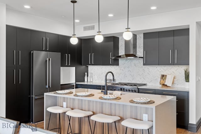 kitchen with appliances with stainless steel finishes, hanging light fixtures, a breakfast bar, a kitchen island with sink, and wall chimney range hood