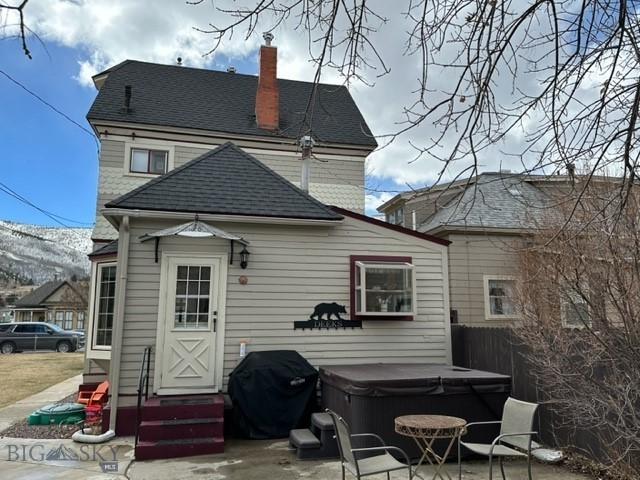 rear view of property featuring entry steps, a patio, a shingled roof, a chimney, and a hot tub