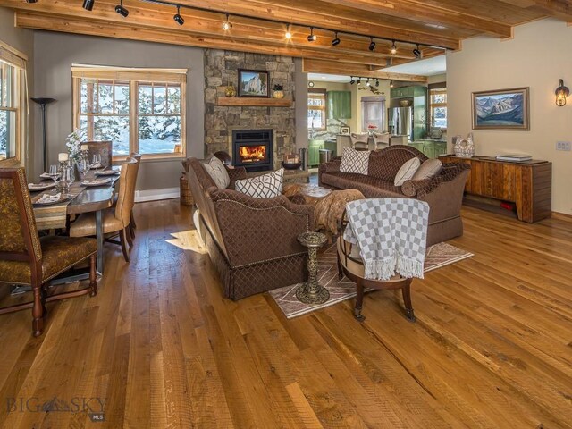 living room featuring a stone fireplace, beamed ceiling, wood-type flooring, and track lighting